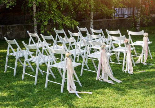 Beautiful wedding set up. Wedding ceremony in the garden. White wooden chairs decorated with flowers and pink ribbons standing in rows. wedding rental, party rental